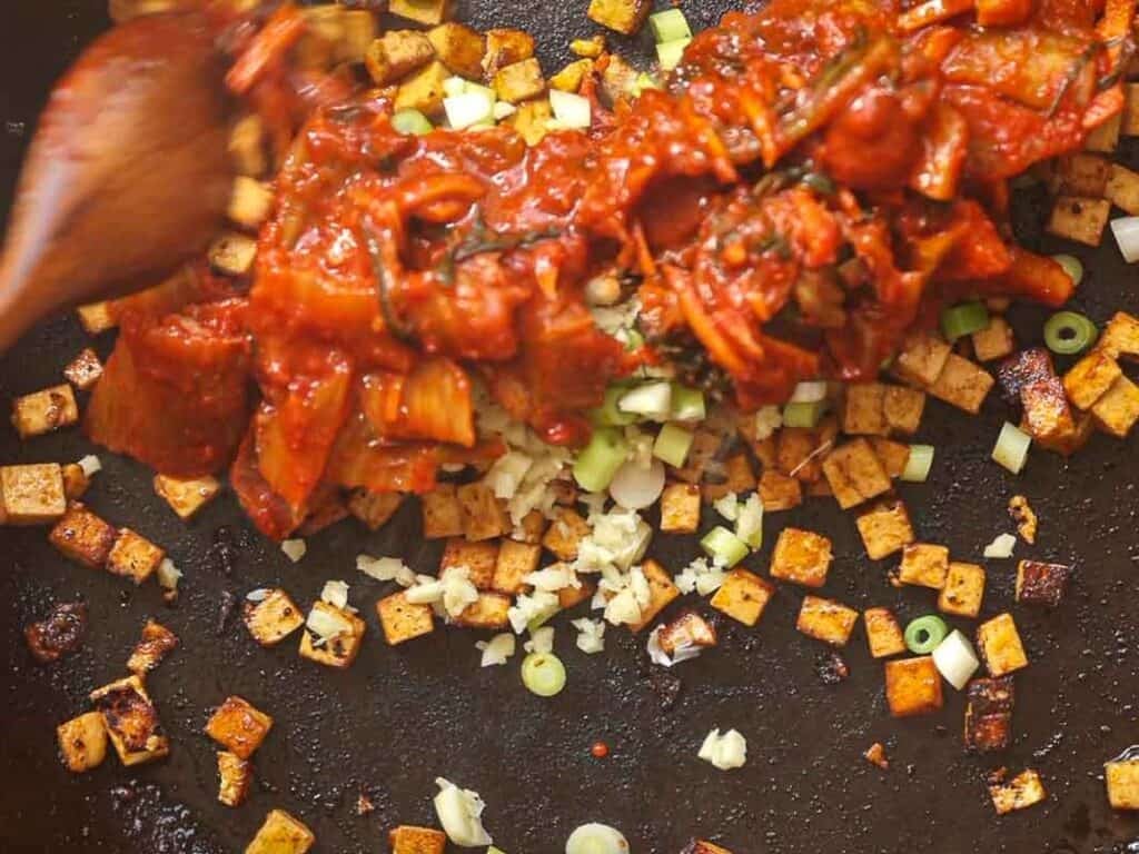 A skillet with sizzling tofu cubes being mixed with spicy kimchi, chopped garlic, and spring onions using a wooden spoon. The tofu is golden brown, and the vibrant red kimchi is spread across the top.